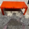 Une table basse « 1970 » d'un orange vif, au design rectangulaire, trône sur une terrasse en pierre, baignée à la fois de soleil et d'ombre. Brocante 93