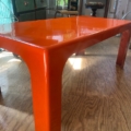Une table basse « Table basse 1970 » orange, moderne et brillante, aux pieds incurvés, trône sur un parquet dans une pièce baignée de soleil. Brocante 93