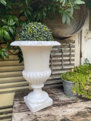 Un vase Médicis blanc du XXe siècle, orné d'un feuillage verdoyant, trône sur une table rustique en bois, à côté d'un pot métallique rempli de plantes, évoquant l'élégance intemporelle du design du début du XXe siècle. Brocante 93