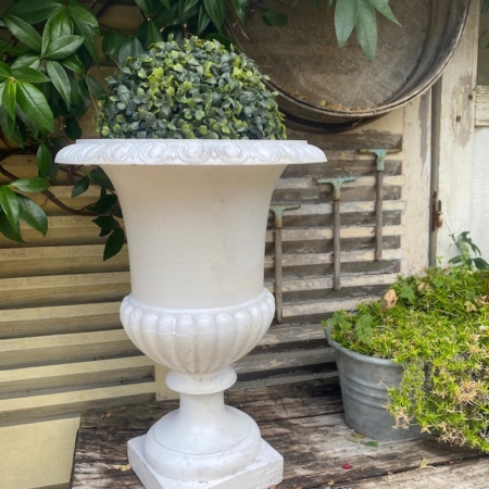 Un vase Médicis blanc du XXe siècle, orné d'un feuillage verdoyant, trône sur une table rustique en bois, à côté d'un pot métallique rempli de plantes, évoquant l'élégance intemporelle du design du début du XXe siècle. Brocante 93