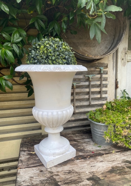 Un vase Médicis blanc du XXe siècle, orné d'un feuillage verdoyant, trône sur une table rustique en bois, à côté d'un pot métallique rempli de plantes, évoquant l'élégance intemporelle du design du début du XXe siècle. Brocante 93