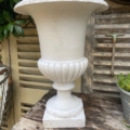 Le vase Médicis blanc du XXe siècle, orné de délicats détails cannelés, est exposé sur une table en bois patiné à l'extérieur, au milieu des volets et des plantes ; cette urne blanche raffinée incarne l'élégance intemporelle du XXe siècle. Brocante 93