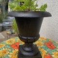 Un vase Médicis du XXe siècle orné de feuilles vertes trône sur une nappe à motifs floraux aux couleurs vives, à l'extérieur. Brocante 93