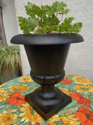 Vase Médicis du XXe siècle, de couleur noire, mis en valeur avec des feuilles vertes sur une nappe à motifs de tournesols, devant un mur clair. Brocante 93