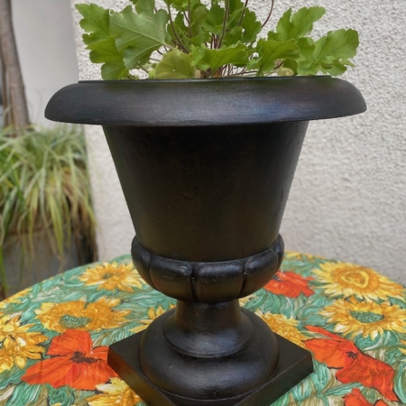 Vase Médicis du XXe siècle, de couleur noire, mis en valeur avec des feuilles vertes sur une nappe à motifs de tournesols, devant un mur clair. Brocante 93