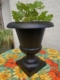 Vase Médicis du XXe siècle, de couleur noire, mis en valeur avec des feuilles vertes sur une nappe à motifs de tournesols, devant un mur clair. Brocante 93