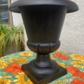 Un vase Médicis du XXe siècle, orné d'un grand motif noir, est posé sur une nappe à fleurs, devant un mur texturé avec des plantes en arrière-plan. Brocante 93