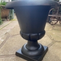 Vase Médicis du XXe siècle, urne-jardinière en métal noir à large rebord, installée sur une terrasse, entourée de plantes et de mobilier d'extérieur. Brocante 93