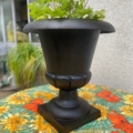 Vase Médicis du XXe siècle orné de feuilles vertes, posé sur une nappe à motifs floraux colorés, dans un décor de plein air inspiré du XXe siècle. Brocante 93