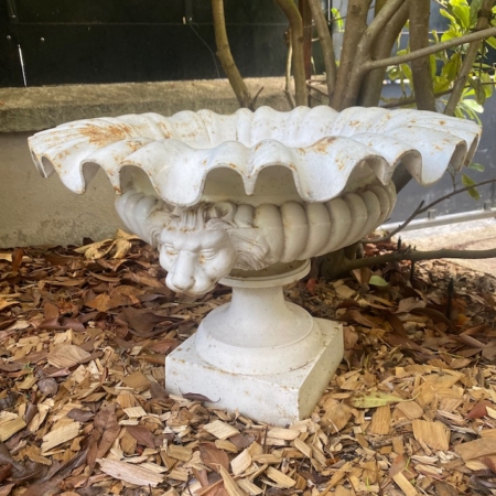 La vasque « Médicis XXe siècle », une jardinière décorative blanche ornée de têtes de lions, est posée sur du paillis sous un buisson feuillu dans le jardin. Brocante 93