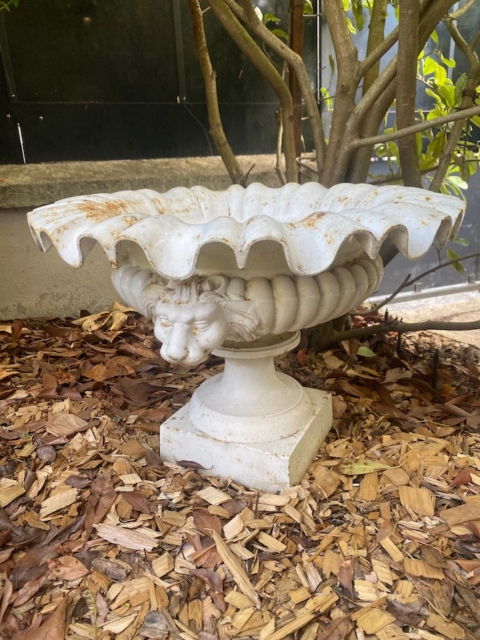 La vasque « Médicis XXe siècle », une jardinière décorative blanche ornée de têtes de lions, est posée sur du paillis sous un buisson feuillu dans le jardin. Brocante 93