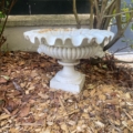 Une vasque Médicis du XXe siècle, une jardinière décorative blanche, repose vide sur des copeaux de bois sous un arbre, près d'un mur en béton. Brocante 93