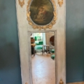 Un miroir trumeau du XIXe siècle, orné d'un tableau ovale, de moulures dorées et de motifs représentant des urnes, est appuyé contre un mur bleu. Brocante 93