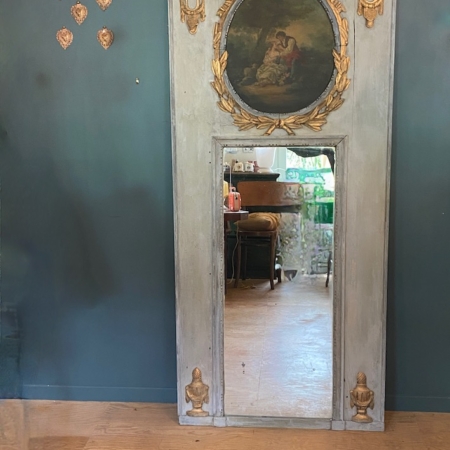 Le miroir trumeau du XIXe siècle, avec son haut cadre ancien et ses touches dorées, se détache sur un mur bleu, entouré de décorations murales, reflétant ainsi le décor éclectique d'une pièce d'inspiration XIXe siècle. Brocante 93