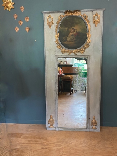 Le miroir trumeau du XIXe siècle, avec son haut cadre ancien et ses touches dorées, se détache sur un mur bleu, entouré de décorations murales, reflétant ainsi le décor éclectique d'une pièce d'inspiration XIXe siècle. Brocante 93