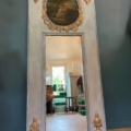 Miroir trumeau du XIXe siècle, orné de motifs dorés raffinés et d’un tableau circulaire dans sa partie supérieure, reflétant un intérieur baigné de soleil. Brocante 93