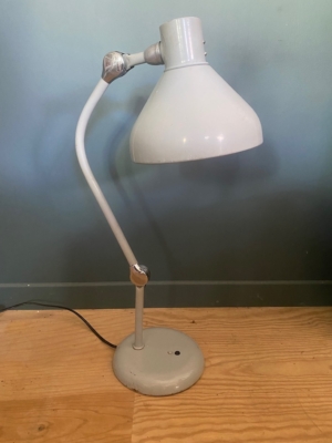 Une lampe de bureau réglable Lampe Jumo GS1 blanche datant de 1950 trône sur un parquet, devant un mur bleu clair, mettant en valeur le design intemporel des années 1950. Brocante 93