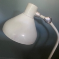 Une lampe de bureau Lampe Jumo GS1 de 1950, de couleur blanche, dotée d'un bras incurvé et d'un design emblématique du milieu du siècle, est posée sur une surface en bois, devant un mur vert uni. Brocante 93