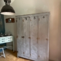 Un meuble « Vestiaire en bois 1940 » doté de quatre portes numérotées trône à côté d'un bureau, sous une suspension industrielle, apportant une touche de charme vintage qui rappelle le style français du milieu du siècle dernier. Brocante 93