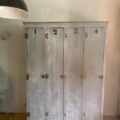Une armoire vintage grise « Vestiaire en bois 1940 », dotée de quatre portes numérotées, trône sur un parquet, adossée à un mur clair. Brocante 93
