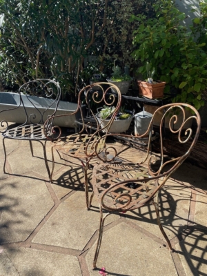 Trois chaises en fer forgé de 1940, richement décorées, trônent sur une terrasse ensoleillée, entourées de plantes verdoyantes et de succulentes en pot, évoquant l'élégance intemporelle du mobilier vintage. Brocante 93