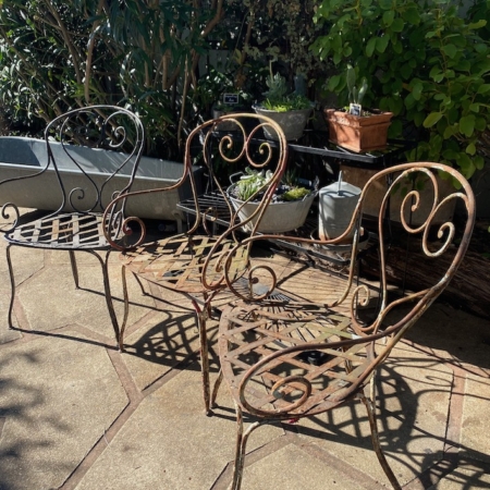 Trois chaises en fer forgé de 1940, richement décorées, trônent sur une terrasse ensoleillée, entourées de plantes verdoyantes et de succulentes en pot, évoquant l'élégance intemporelle du mobilier vintage. Brocante 93
