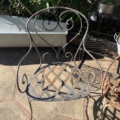 Une chaise en fer forgé datant de 1940, dotée d'une assise en treillis et d'un dossier incurvé, trône sur une terrasse en pierre baignée de soleil. Brocante 93