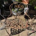 Des chaises en fer forgé des années 1940, dotées d'une assise en treillis, trônent dans un jardin ensoleillé, entourées de plantes grasses en pot, évoquant le charme intemporel du design français d'époque. Brocante 93