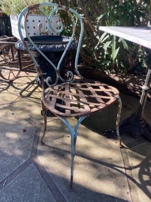 Installée sur une terrasse ensoleillée, près d'une table et de buissons verdoyants, la chaise en fer forgé de 1940 — avec son cadre en métal rouillé et son assise en treillis — évoque le charme intemporel du mobilier vintage des années 1940. Brocante 93