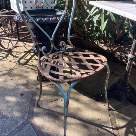 Installée sur une terrasse ensoleillée, près d'une table et de buissons verdoyants, la chaise en fer forgé de 1940 — avec son cadre en métal rouillé et son assise en treillis — évoque le charme intemporel du mobilier vintage des années 1940. Brocante 93
