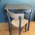 Un bureau d’enfant et une chaise « 1960 » gris et beige se détachent sur un mur bleu sarcelle, posés sur un parquet. Brocante 93