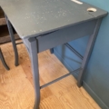 Le bureau d’enfant et sa chaise datant des années 1960, avec son bois gris vieilli et son plateau usé orné d’un trou pour l’encrier et d’une rainure pour les crayons, trône sur un parquet à côté d’un mur bleu, conférant à la scène un charme nostalgique. Brocante 93
