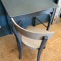 Un bureau d’enfant gris datant des années 1960, accompagné d’une chaise, trône sur un parquet près d’un mur bleu. Brocante 93