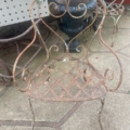 Chaises en fer forgé datant de 1940, ornées de volutes élaborées sur le dossier et présentant une patine rustique, disposées sur une terrasse en pierre. Brocante 93