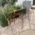Deux chaises en fer forgé datant de 1940 sont disposées sur une terrasse en pierre ; leurs structures métalliques d'époque sont entourées de plantes vertes en pot. Brocante 93