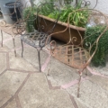 Deux chaises en fer forgé de 1940, richement décorées et patinées par les intempéries, sont disposées à côté d'une jardinière remplie de plantes retombantes vertes, sur une terrasse en pierre. Brocante 93