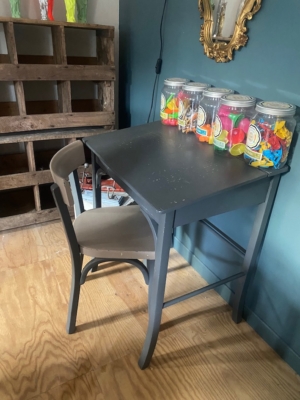 Un bureau d’enfant et une chaise datant des années 1960, ornés de bocaux remplis de ballons colorés, sont posés sur un parquet, devant un mur vert. Brocante 93