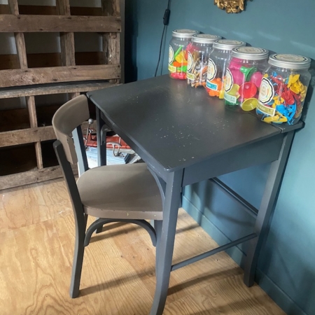 Un bureau d’enfant et une chaise datant des années 1960, ornés de bocaux remplis de ballons colorés, sont posés sur un parquet, devant un mur vert. Brocante 93