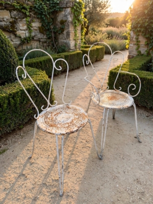 Deux chaises en fer forgé de 1950, en métal blanc, avec des dossiers en forme de cœur, disposées le long d'une allée de jardin. Brocante 93