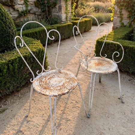 Deux chaises en fer forgé de 1950, en métal blanc, avec des dossiers en forme de cœur, disposées le long d'une allée de jardin. Brocante 93