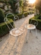 Deux chaises en fer forgé de 1950, en métal blanc, avec des dossiers en forme de cœur, disposées le long d'une allée de jardin. Brocante 93