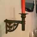 Une bougie rouge dans une applique murale en laiton « Chandelier de piano 1960 » ornée d'un motif floral, sur un mur clair. Brocante 93